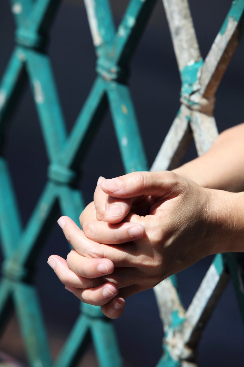 A close-up shot of clasped hands placed through prison bars. Image: iStock