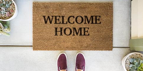 Woman standing in front of a doormat reading “It’s so good to be home”.