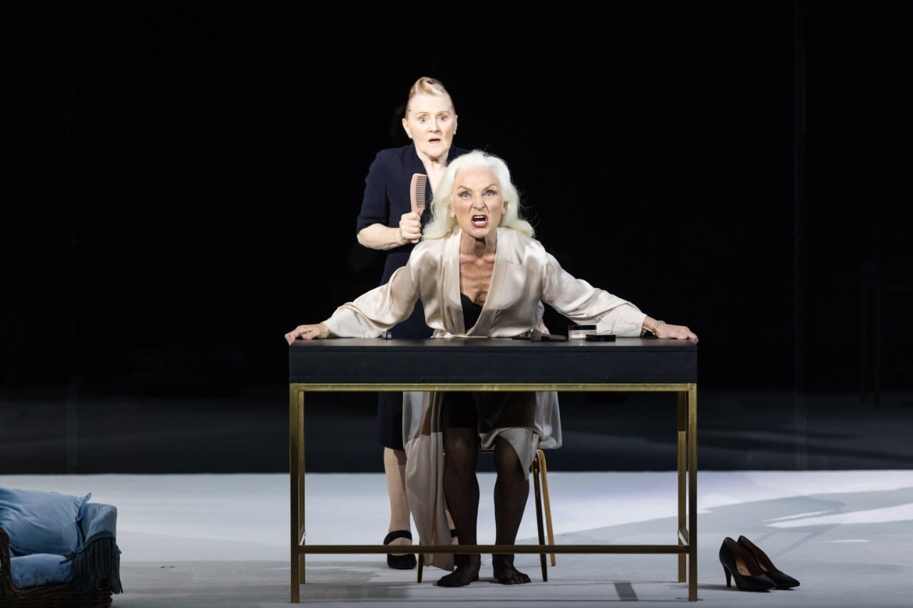 two female actors on a stage, one is sitting behind a table and gripping it and pulling an angry face as though she is shouting