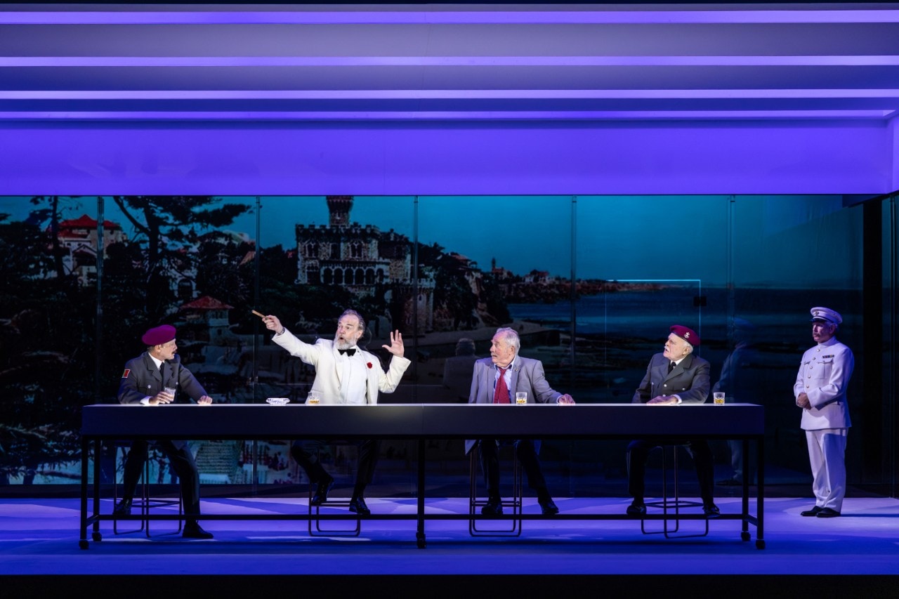 a group of male actors are sitting at a long dining table, one of them is shouting and pointing