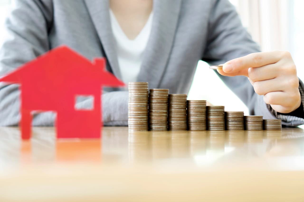 A series of stacked coins on a table beside a graphic of a house, indicating rising household debt. Image: iStock