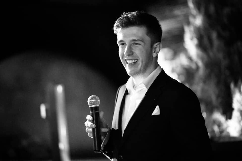 Cooper Sheather in a tuxedo holding a microphone
