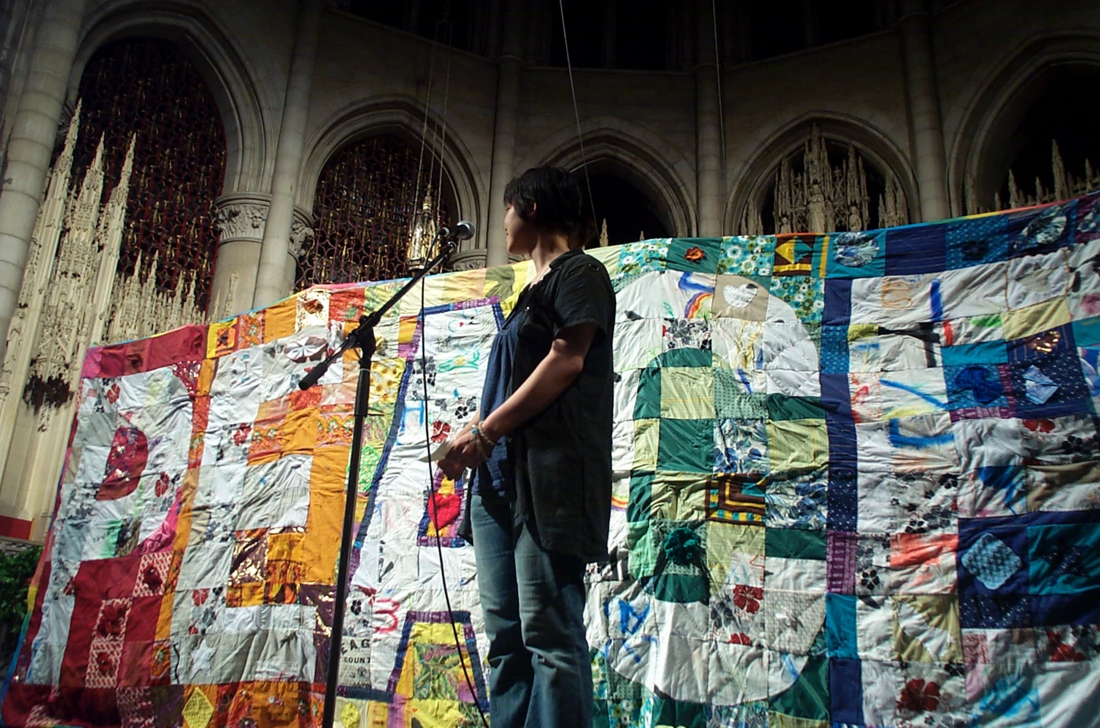 A person with a microphone standing in front of a quilt that spells out 'peace'