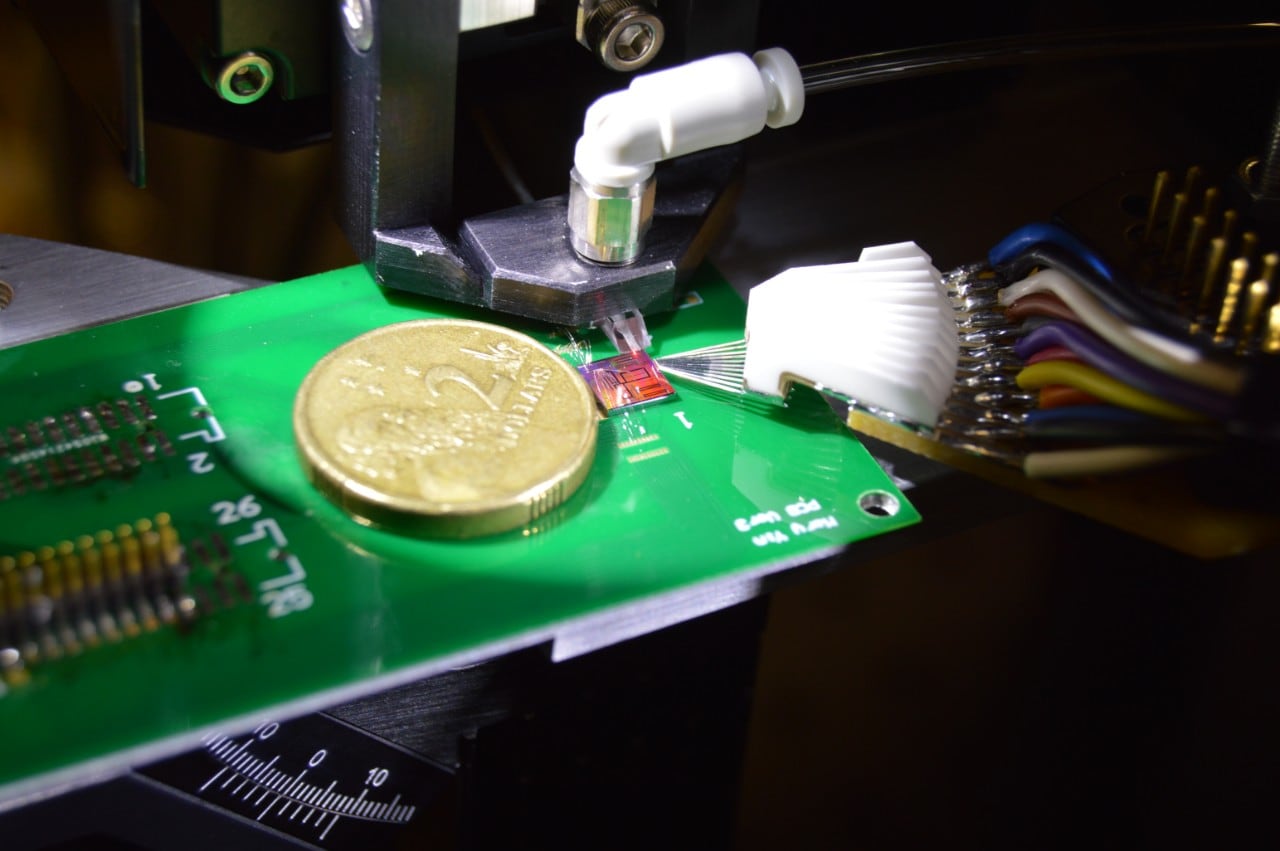A close up image of a microchip with an australian two dollar coin next to it to show the microchip is about one tenth the size of the coin. The microship is on a green microchip board with many sensors poitning towards the chip.