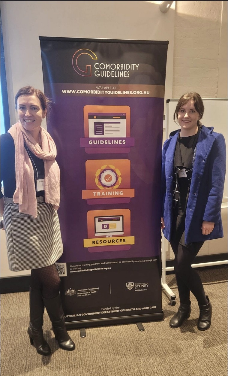 Two women wearing coats standing next to a large banner that says 'the comorbidity guidelines'