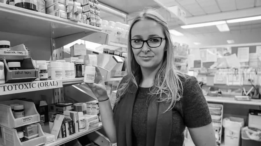 Black and white image of pharmacist Rose Cairns standing at shelves of medicines holding a small bottle
