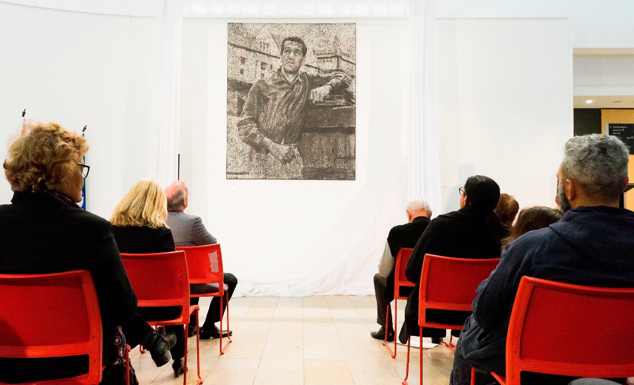 A photo of people seated, looking at a large black and white dot painting of Charles Perkins.