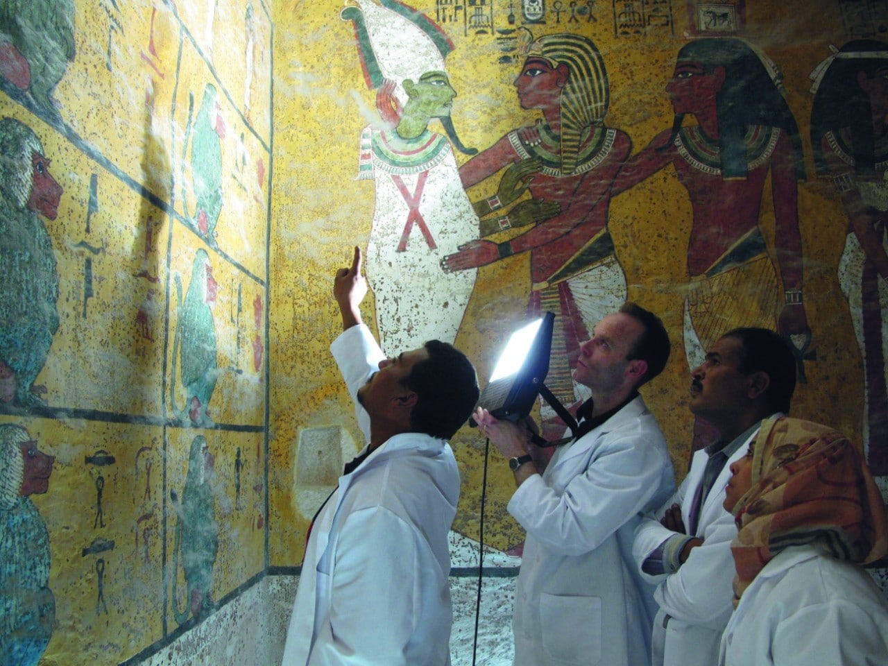 Four people in white lab coats - one pointing a large light at the wall - are looking at decoratively painted walls in the tomb of Tutankhamen.