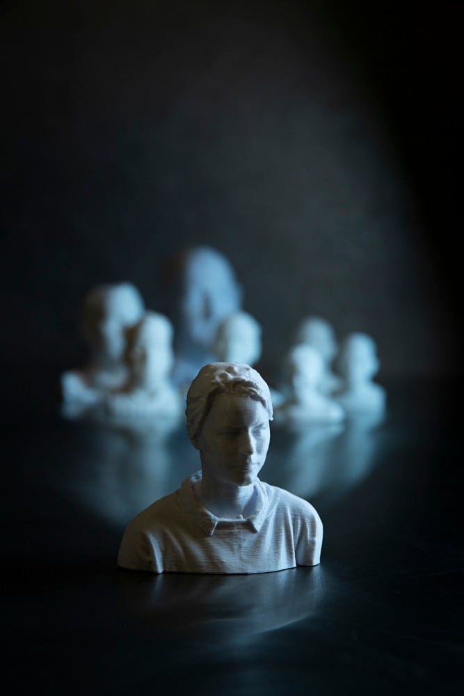 Sitting on a polished table is a 3D printed, head and shoulders rendering of Cheng. It is like white marble, and she is wearing a collared shirt. Behind her is a crowd of 3D models of Cheng's workmates, though they are blurry and indistinct. The image has a darkness about it but side lit in tones of blue.