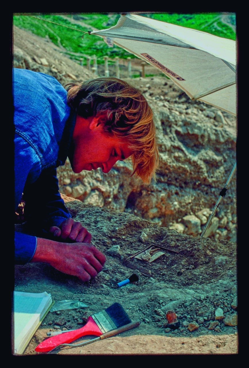A young Tim Potts in Pella, Jordan, blonde hair tumbling forward, he is hunched over a small object - the lion box - which he is carefully removing from the grey earth and rubble that is holding it. Beside him is a large bright red paint brush, that he has been using to sweep dirt away.