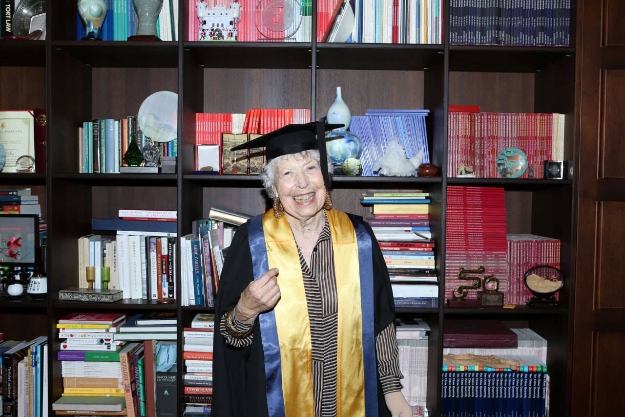 Older woman in academic gown in front of a book casing smiling