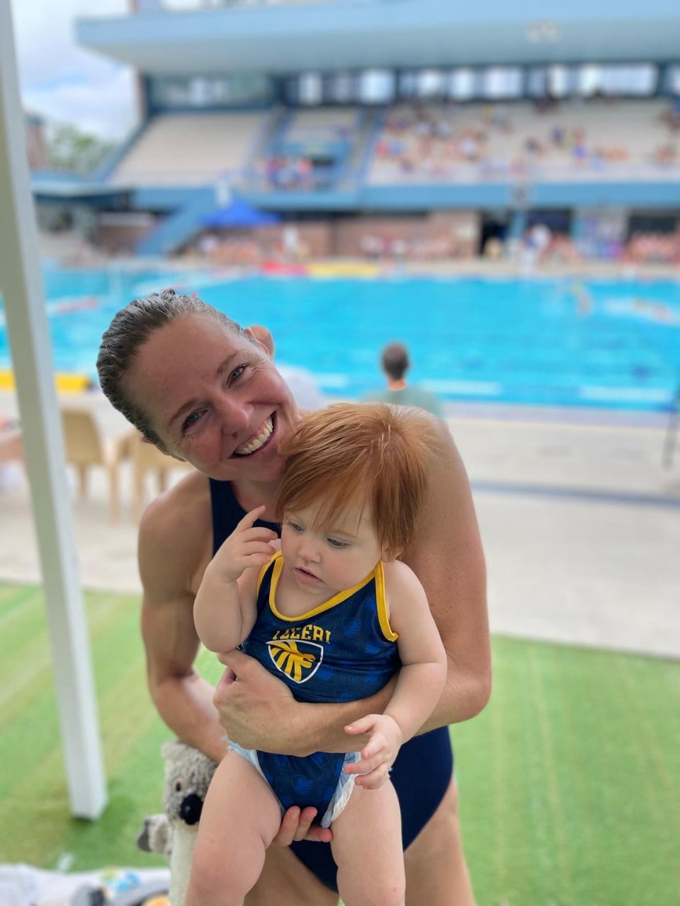 A woman in a swimming costume holds a baby beside a swimming pool.