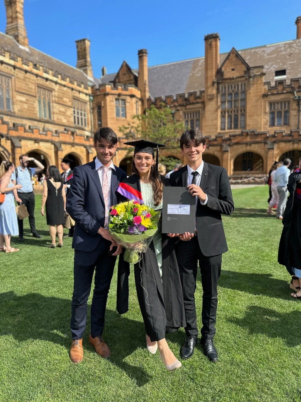 A young woman in graduation robes stands between two young men in suits outside a sandstone building.