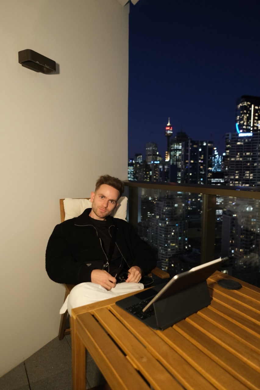 Pasha sitting on a table by his balcony with a laptop in front of him, the background overlooks the city night sky.
