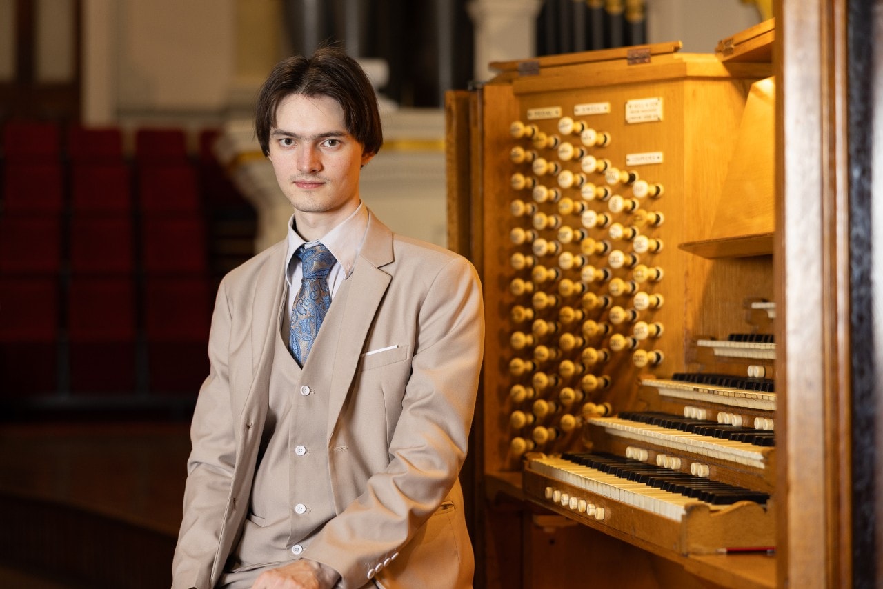 Titus Grenyer seated at the University organ, facing the camera and smiling. He is wearing a beige three-piece suite with a blue tie. 