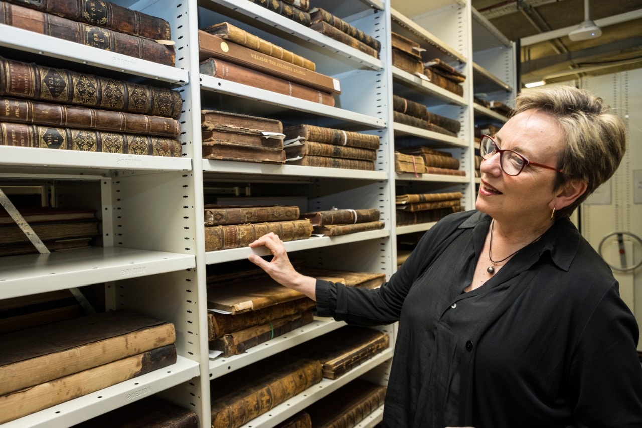 Professor Louise Baur taking a look at Fisher Library's Rare Books and Special Collections. Shelves of books can be seen in front of her.
