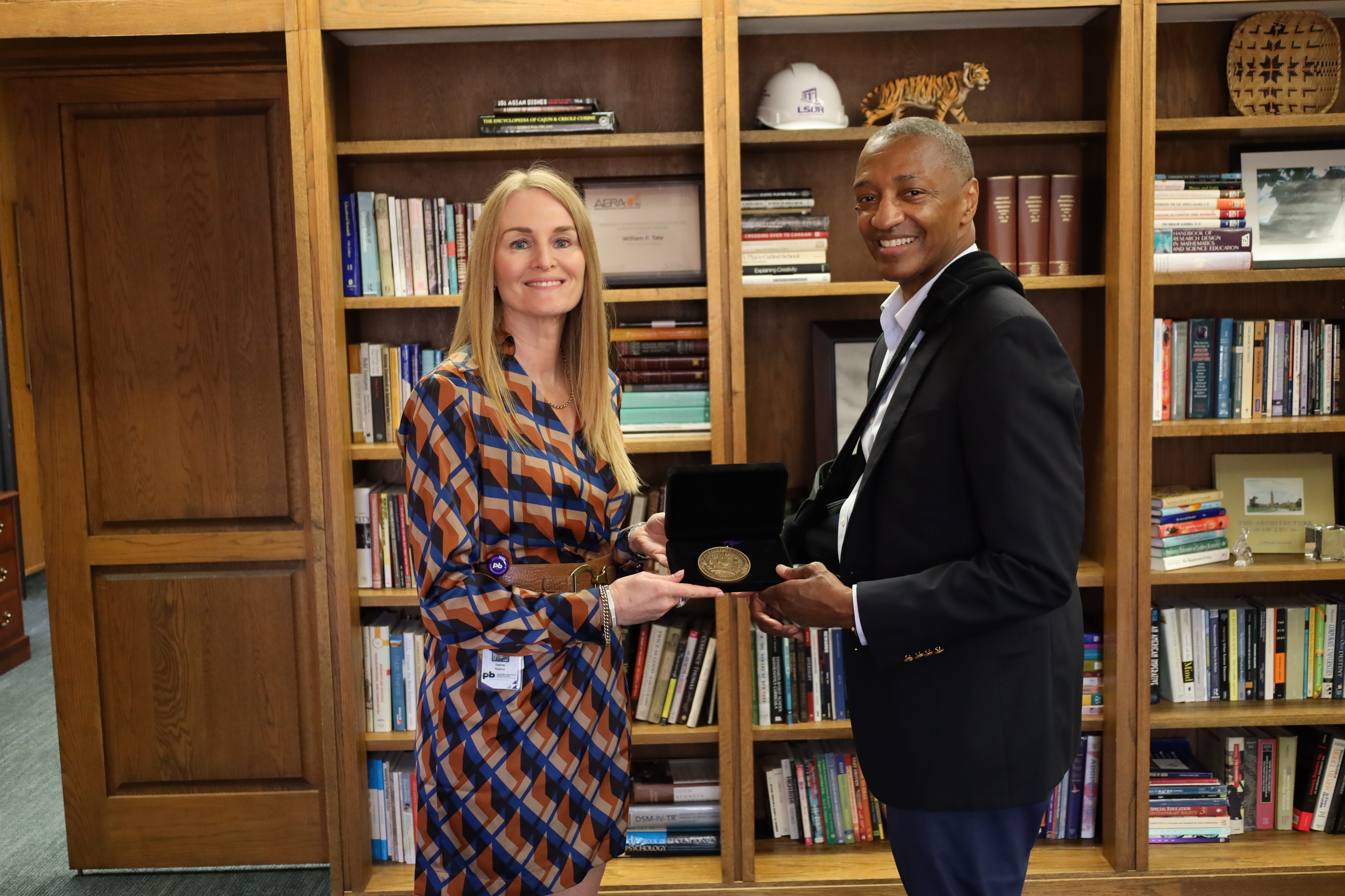 Professor Leanne Redman receives a medal from Louisiana State University President William Tate. Redman and Tate are holding the medal and smiling at the camera, standing against a wall lined with bookcases.