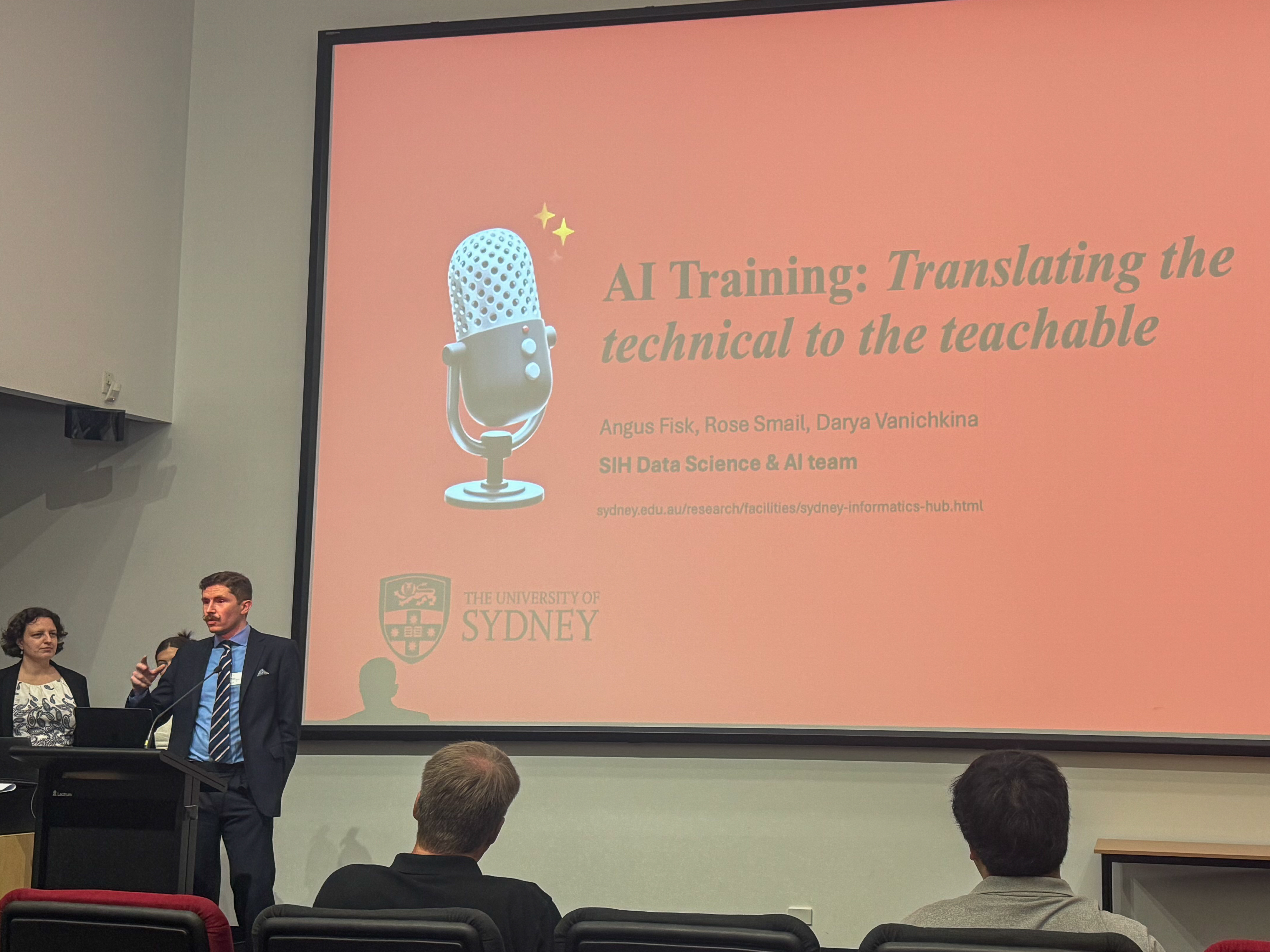 Dr Angus Fisk presenting at a conference. The slideshow projected behind him reads