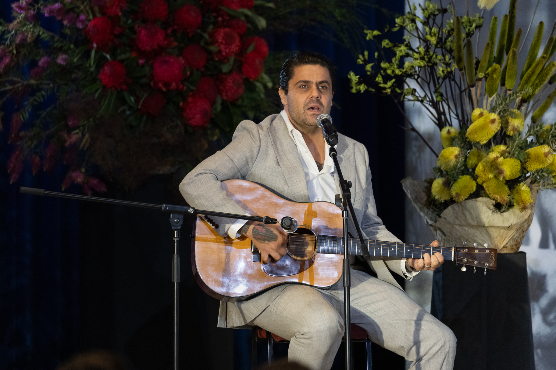 A man is seated playing guitar on a stage with flowers behind him.