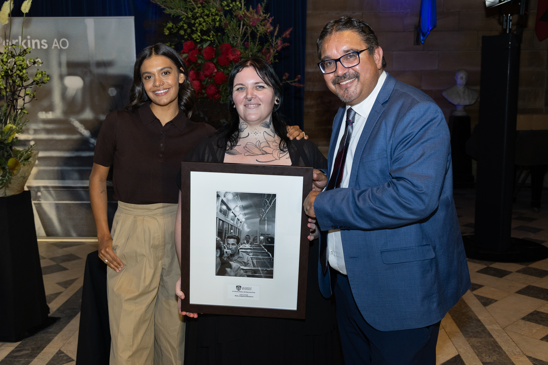 Three people smiling for a photo, two women and a man on the right. The woman in the centre holds a picture frame. 