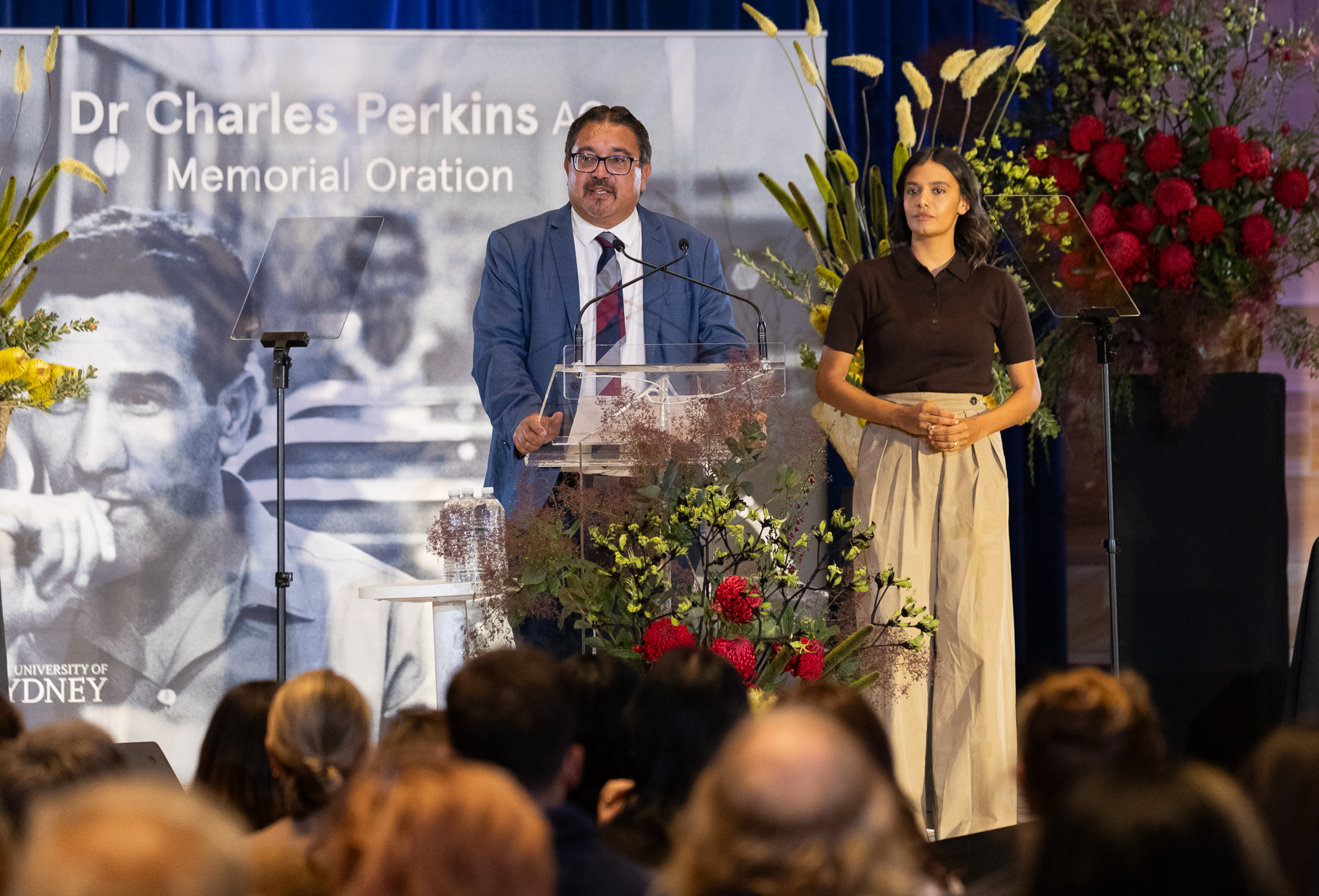 A man standing at a podium with a woman next to him, with a media wall and floral work.