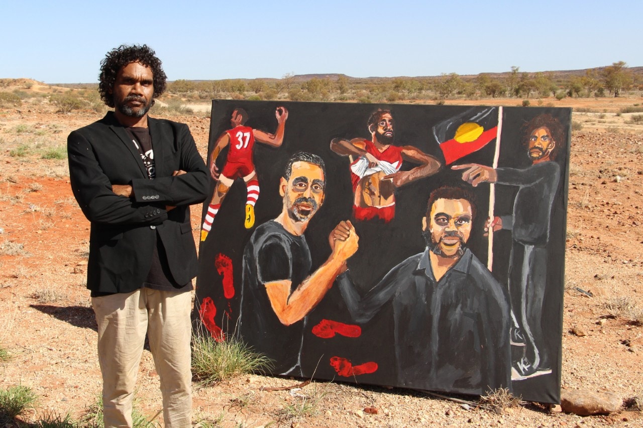 Australian indigenous artist Vincent Namatjira in the desert standing next to his painting of footballer Adam Goodes
