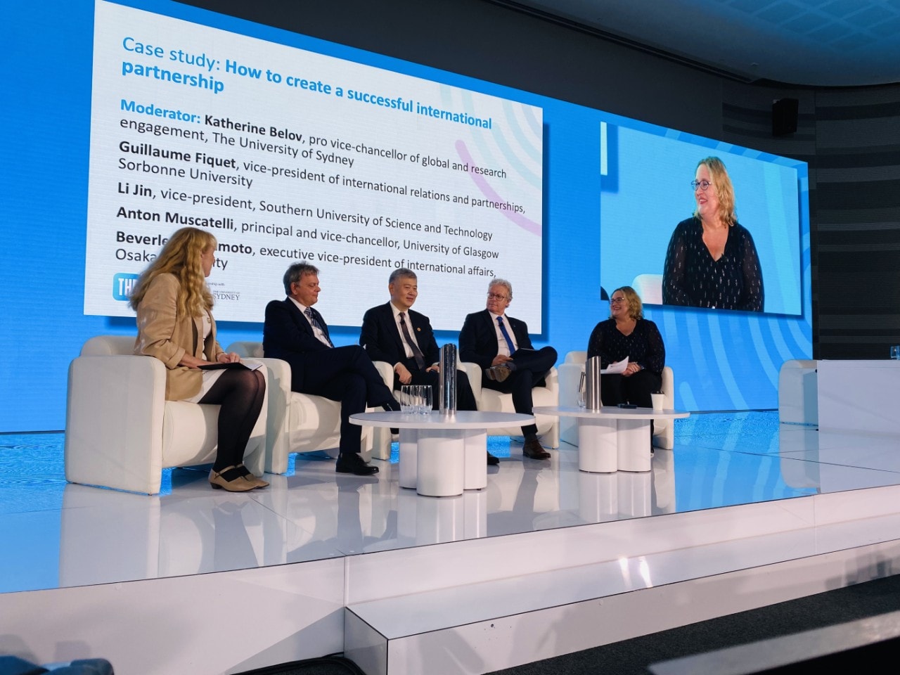 Five panellists seated on stage with screen displayed behind them