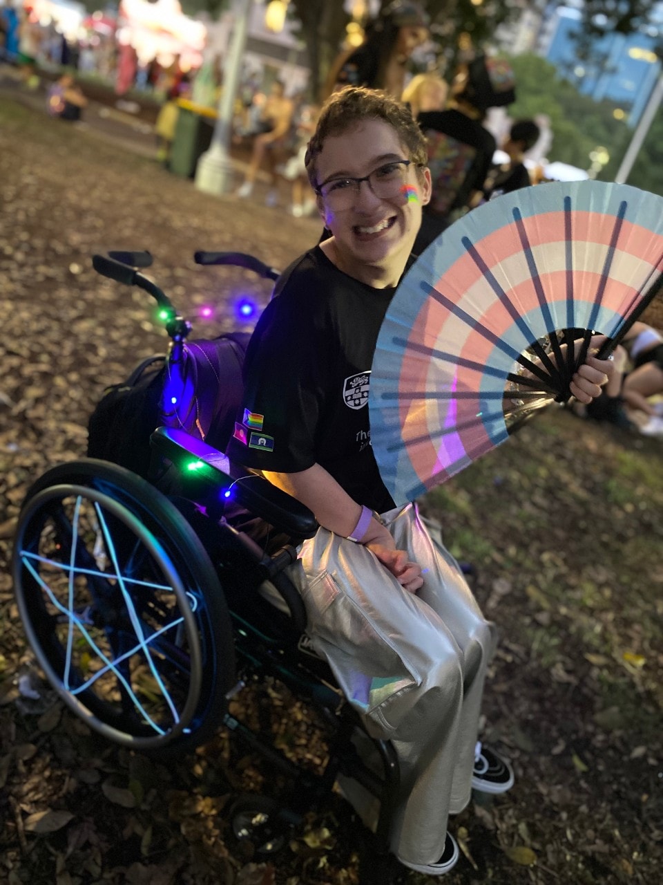 Riley sitting in wheelchair decorated with brightly coloured neon lights, holding a fan featuring the Transgender Pride Flag