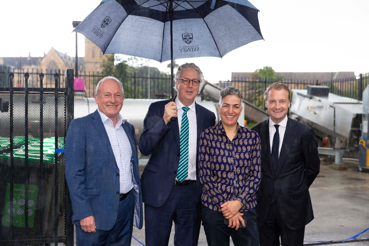 A group of four people standing in front of a machine in the rain, one holds an umbrella