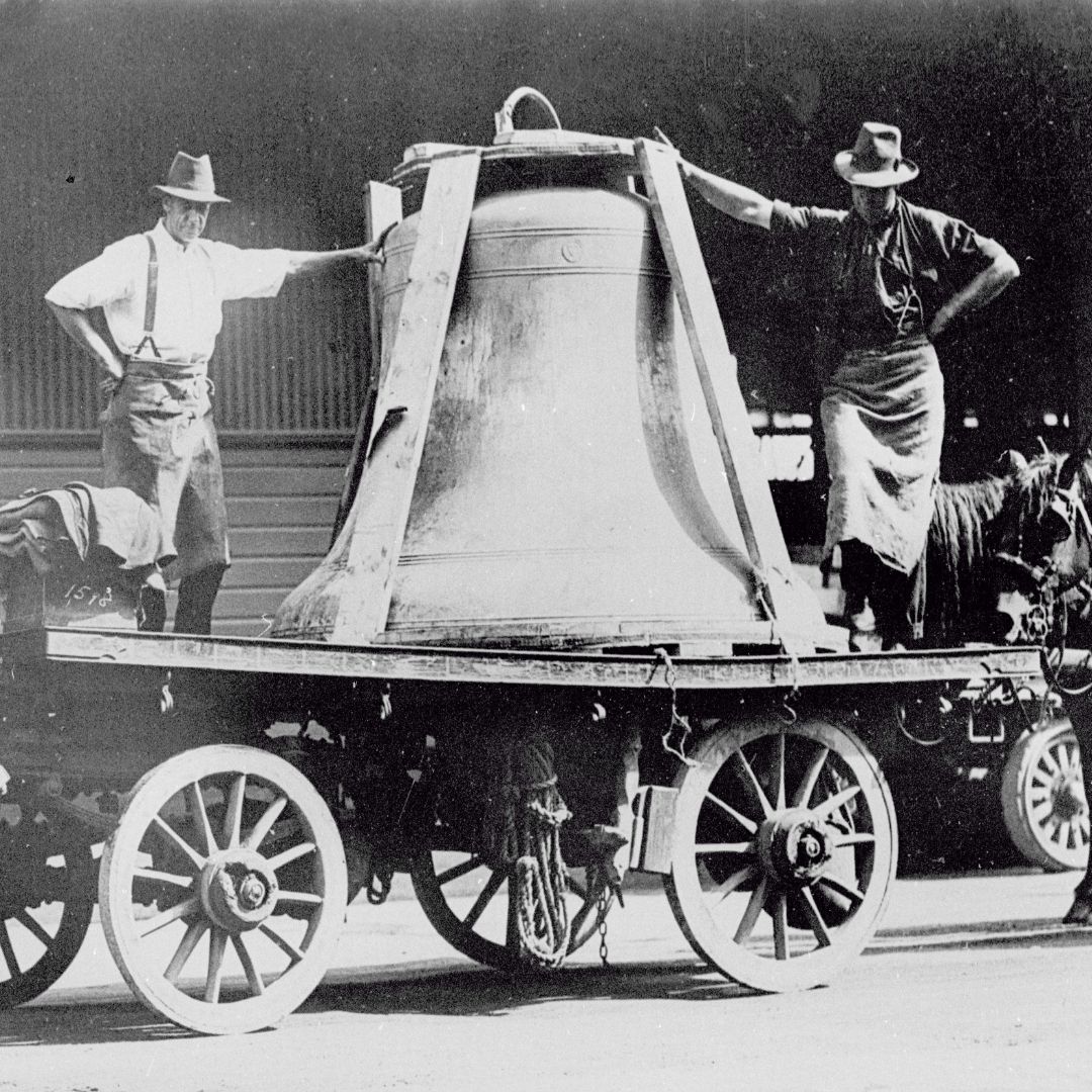 Two men standing on top of a carriage near the large bell in a black and white archival image.