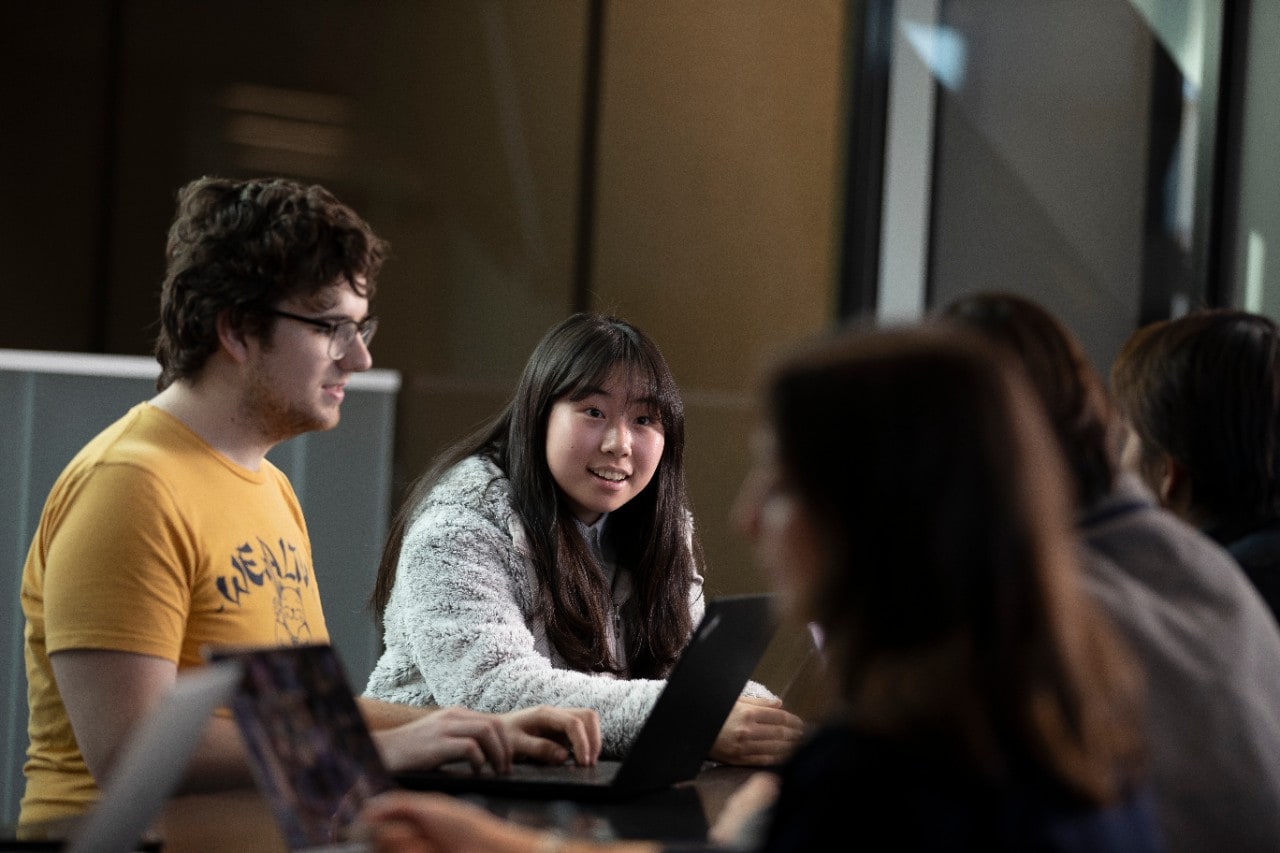 A group of students smiling and sitting around a table with laptops