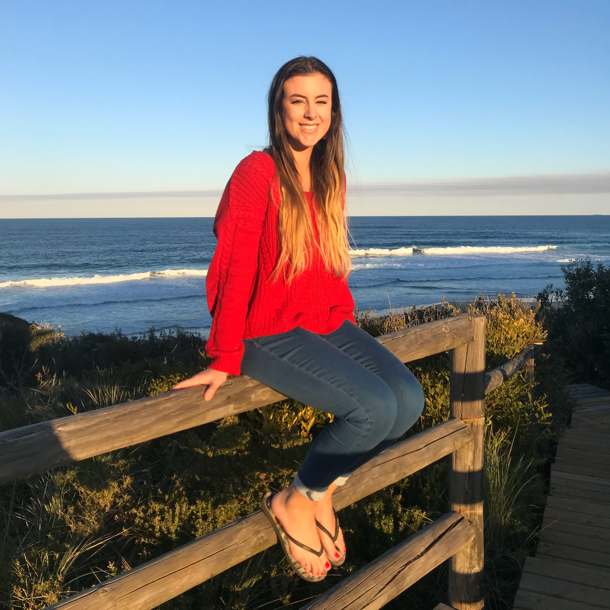 Maddi sits on a fence with the beach behind her on the South Coast of NSW.