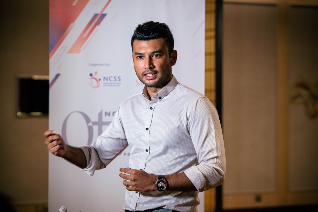 Dr Vimallan Manokara wearing a white business shirt and standing in front of a banner while presenting to an audience