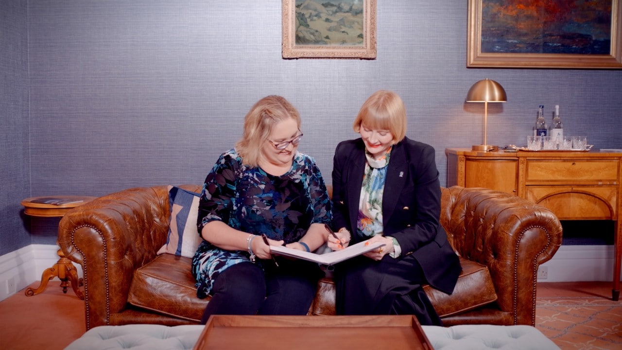 A photo of Professor Kathy Belov and Rachel Sandison, sitting on a lounge signing the new agreement.