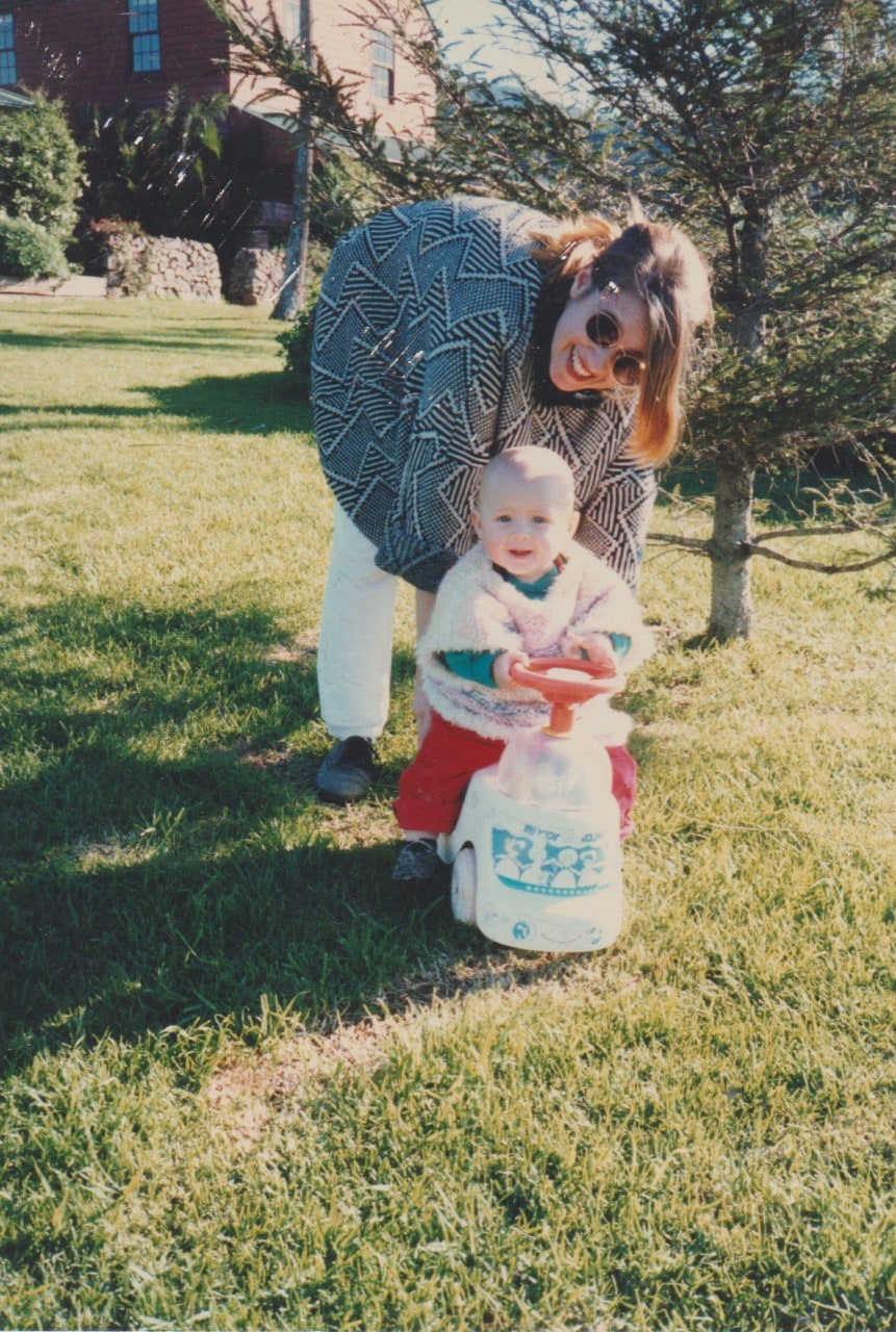 Young Anne Howell leaning over in front of a tree smiling with a baby who is on a scooter.  