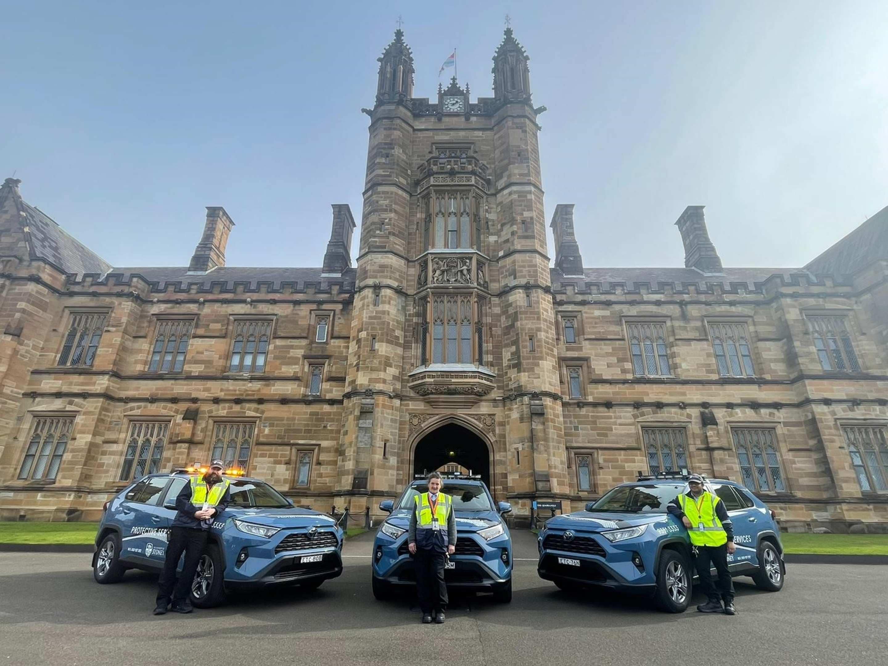 Three blue Protective Services vehicles are parked in front of the Quad. A Protective Services staff member stands in front of each of them wearing yellow high-visibility vests.