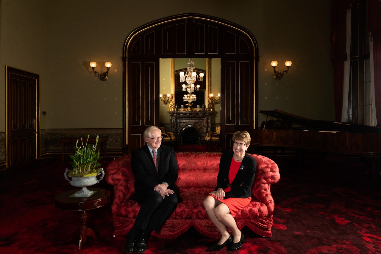 The vice regal couple seated on a plush, old style and bright red sofa. Mr Wilson, on the far left, is wearing a dark business suit. Governor Beazley on the far right, is in a black skirt with a red jacket. Both are smiling warmly. Behind them is a large entrance into another formal area and we can see a mirror reflecting chandeliers.